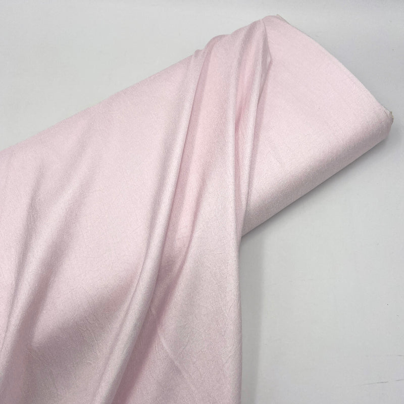 A bolt of light pink fabric with a subtle crepe texture sitting on a plain white table. 