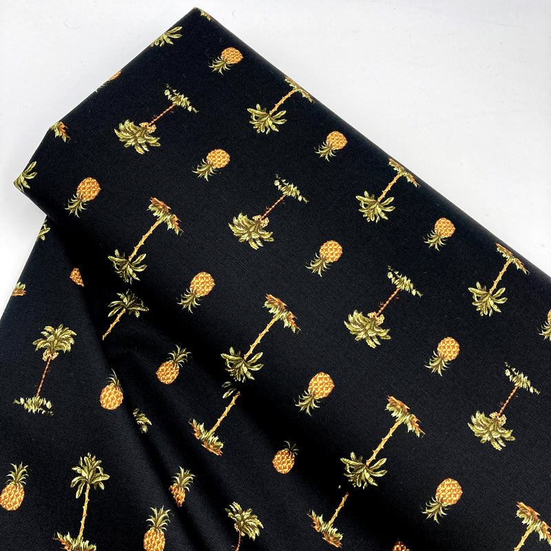A bolt of black fabric with pineapple and palm tree motifs printed on it. The fabric sits on a white background.